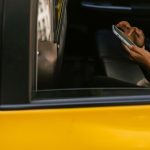 Passageiro usa um smartphone no banco traseiro de um táxi amarelo durante a viagem, com o motorista à frente.