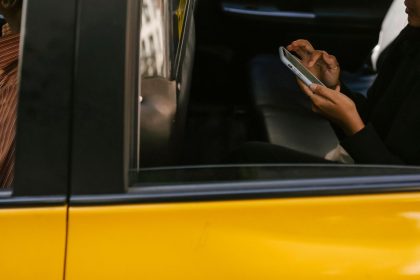 Passageiro usa um smartphone no banco traseiro de um táxi amarelo durante a viagem, com o motorista à frente.