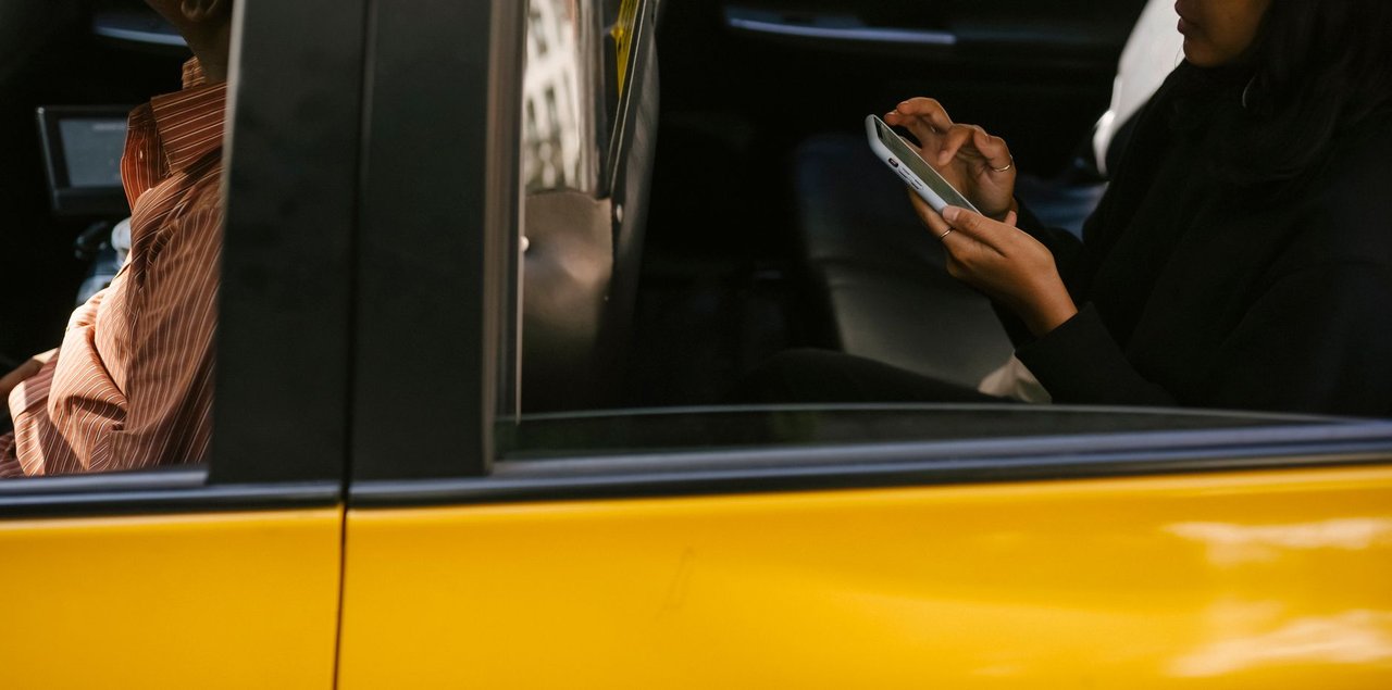 Passageiro usa um smartphone no banco traseiro de um táxi amarelo durante a viagem, com o motorista à frente.
