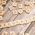Peças de madeira formam a palavra “Regulation” sobre uma superfície texturizada, remetendo à regulamentação de criptoativos.