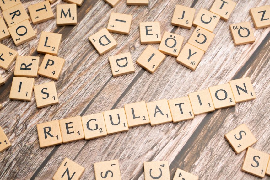 Peças de madeira formam a palavra “Regulation” sobre uma superfície texturizada, remetendo à regulamentação de criptoativos.